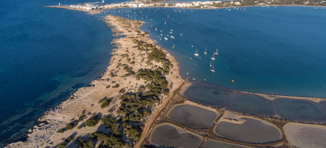 Luftaufnahme von Estany des Peix auf Formentera. Im Vordergrund Salinenbecken, rechts die Bucht mit zahlreichen Segelbooten, dahinter der Ort La Savina