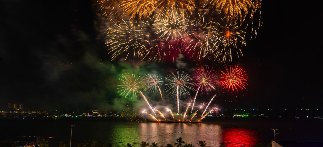 Großes Feuerwerk über dem Meer in Pattaya bei Nacht. Am Strand und auf einer Rooftop-Bar feiern Menschen das farbenfrohe Spektakel.