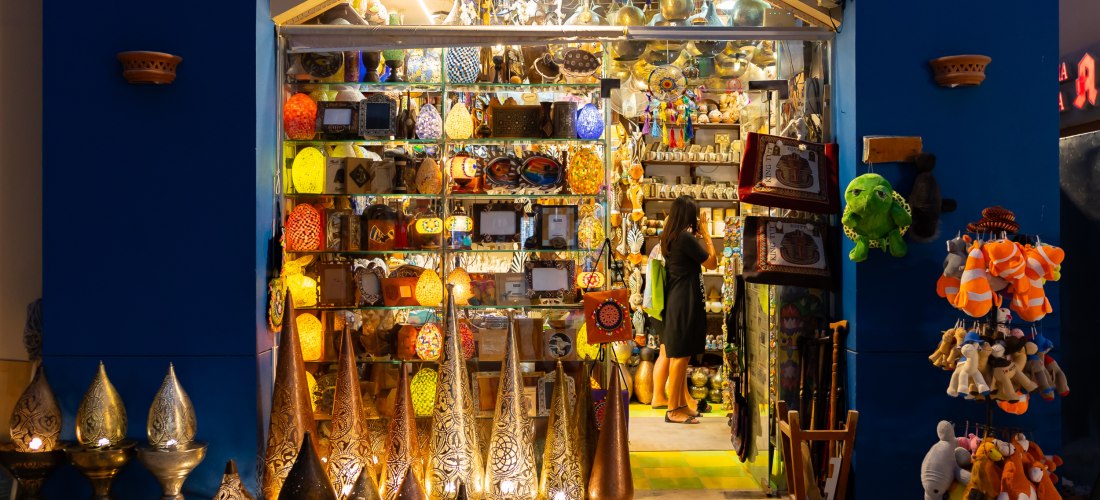 Souvenirshop mit orientalischen Lampen, Keramik, Taschen und Plüschtieren. Eine Frau schaut sich im Laden um. Die Szenerie ist farbenfroh und abends beleuchtet.