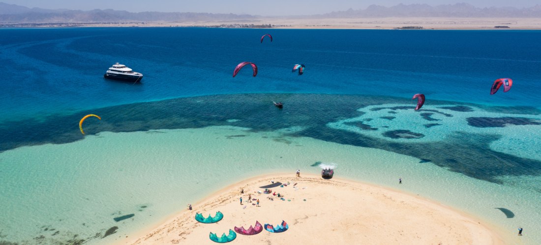 Bunte Kites schweben über türkisfarbenem Wasser vor einer Sandbank im Roten Meer. Auf der Sandbank liegen mehrere Kites und Menschen bereiten sich auf den Wassersport vor. Im Hintergrund ankert ein Boot, dahinter liegt das Festland mit einer kargen Küsten