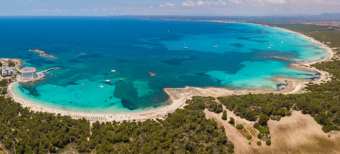 Panoramablick auf eine Küste mit hellem Sandstrand und türkisfarbenem Wasser. Der Strand ist gesäumt von grünen Pinienwäldern, die sich sanft in die Landschaft einfügen. Im Wasser liegen zahlreiche Boote und Yachten verteilt. Ein großes Hotelgebäude mit P