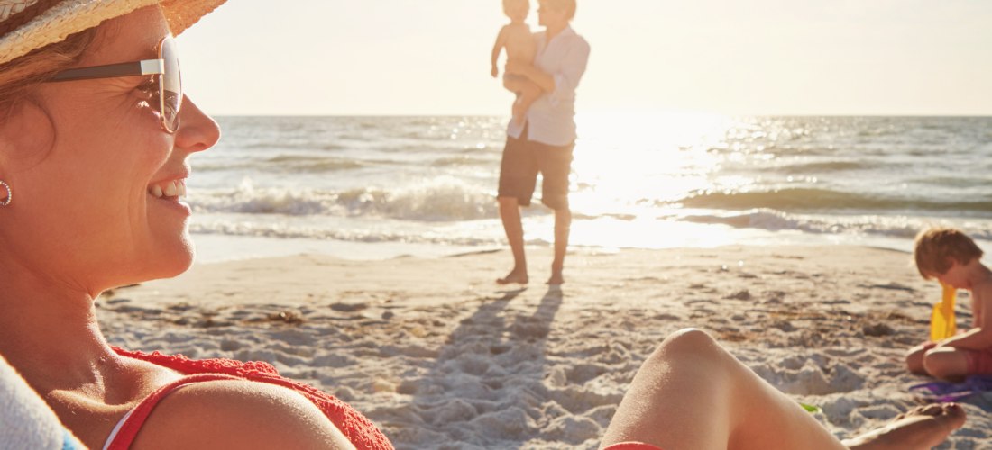 Im Vordergrund liegt eine Frau mit einem Strohhut und Sonnenbrille auf einer bequemen Liege, während die warmen Sonnenstrahlen ihr Gesicht erhellen. Ein gestreiftes Handtuch und eine gelbe Tasche verleihen der Szene eine sommerliche Atmosphäre. Im Hinterg