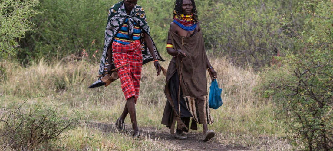 Zwei Angehörige der Samburu-Volksgruppe gehen auf einem schmalen Pfad durch eine bewachsene Savannenlandschaft. Der Mann trägt eine rote karierte Wickelhose, farbige Kleidung und Federschmuck am Kopf, die Frau trägt eine traditionelle braune Robe mit bunt