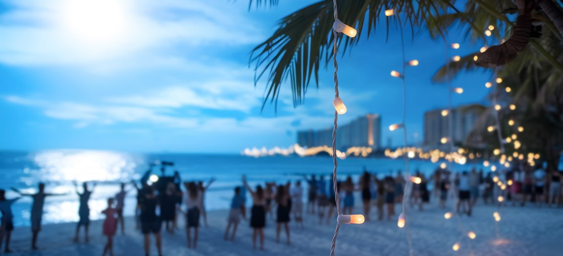 Eine stimmungsvolle Strandparty bei Nacht: Sanft leuchtende Lichterketten hängen an Palmen und erhellen die Szenerie. Im Hintergrund feiern Menschen am Sandstrand unter dem Licht des Mondes, während die Stadtlichter in der Ferne glitzern. Eine perfekte Ku