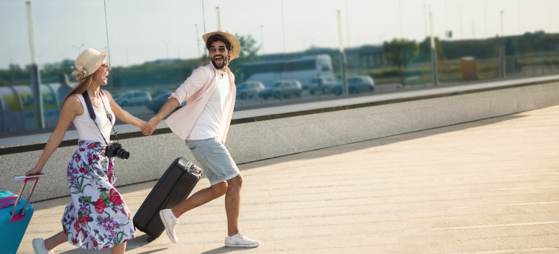 Junges Paar mit Sommerkleidung zieht Rollkoffer und läuft lachend Hand in Hand am Flughafen entlang