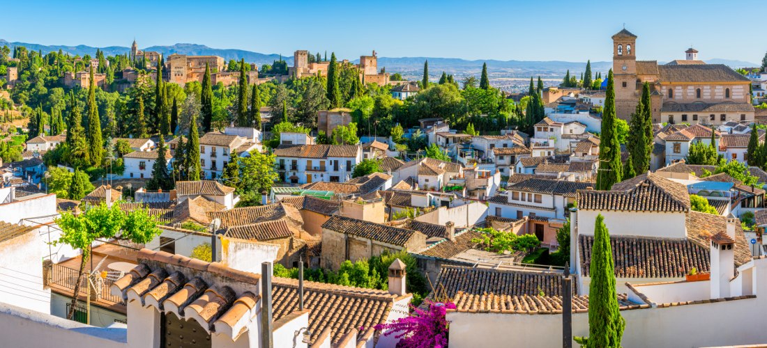 Blick über die Dächer von Granada mit der maurischen Festungsanlage Alhambra auf einem Hügel, umgeben von grüner Vegetation.