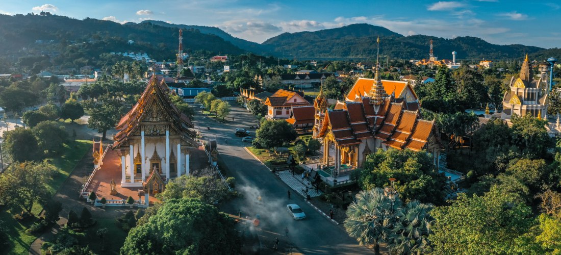 Luftaufnahme einer eindrucksvollen Tempelanlage auf Phuket mit mehreren goldverzierten Gebäuden, roten Dächern und üppigem Grün, eingerahmt von Hügeln und blauem Himmel.