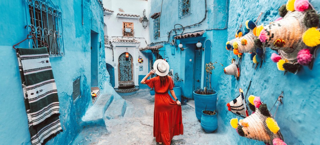 Frau in rotem Kleid spaziert durch eine enge, blau gestrichene Gasse in der Altstadt von Chefchaouen, Marokko – beliebtes Fotomotiv und Sehenswürdigkeit.