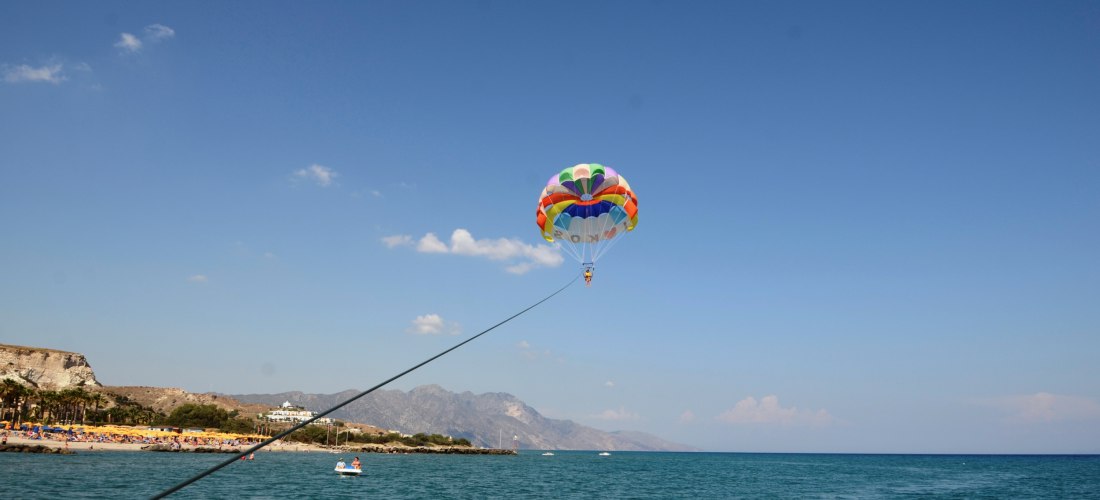 Eine Person schwebt beim Parasailing an einem bunten Fallschirm über dem türkisblauen Meer. Der Fallschirm ist über ein Seil mit einem Boot verbunden, das außerhalb des Bildes liegt. Am linken Bildrand ist ein belebter Sandstrand mit Sonnenschirmen zu seh