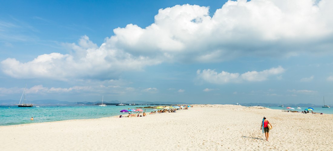 Weißer Sandstrand mit wenigen bunten Sonnenschirmen und Badenden. Eine Person geht mit einem Badetuch über der Schulter in Richtung Wasser.