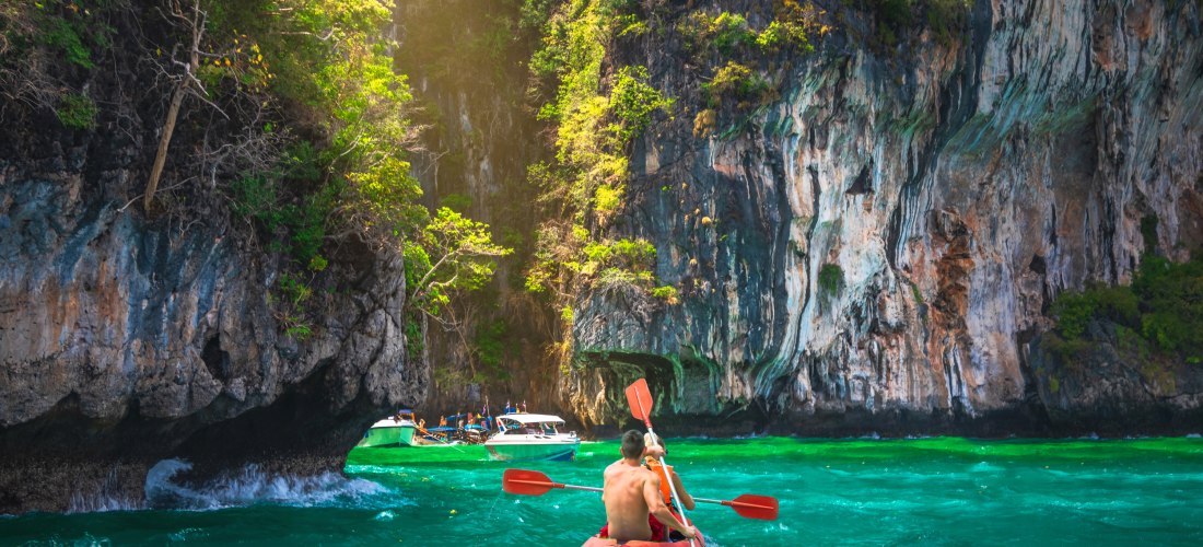 Zwei Personen paddeln in einem roten Kajak durch türkisfarbenes Wasser in einer felsigen Bucht auf Phuket. Umgeben von hohen, mit tropischen Pflanzen bewachsenen Kalksteinfelsen fällt Sonnenlicht dramatisch durch das Blattwerk. Im Hintergrund liegen mehre