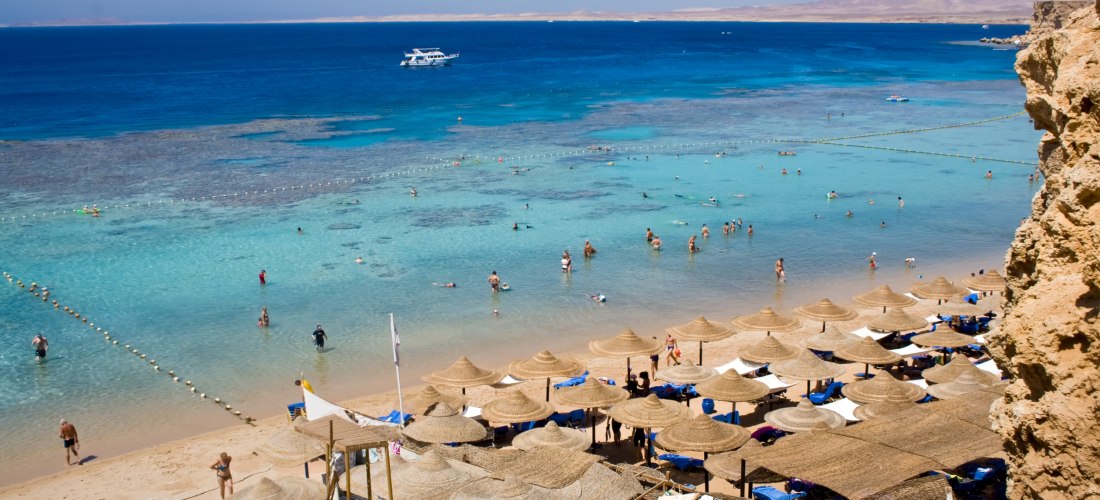 Leute sonnen sich und schwimmen im Meer, am Strand gibt es Strohschirme