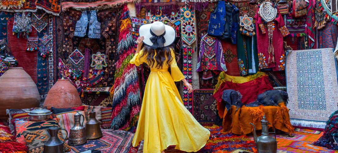 Eine Frau in einem langen, gelben Kleid und mit einem weißen Sonnenhut steht inmitten eines traditionellen orientalischen Markts mit bunten Teppichen, Stoffen, Kleidungsstücken und Kupferwaren. Sie betrachtet die kunstvoll dekorierten Textilien an der Wan