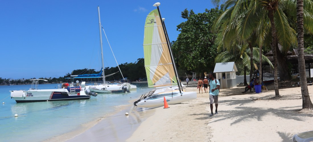 Strand in Jamaika mit Segelbooten und Palmen. Am weißen Sandstrand liegen mehrere Boote im türkisblauen Wasser, darunter ein Katamaran mit farbigem Segel. Eine Person spaziert barfuß am Ufer entlang, während andere unter Palmen im Schatten entspannen. Im 