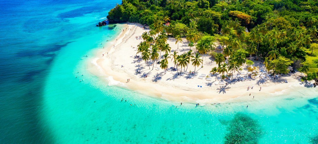 Langer, weißer Sandstrand mit unzähligen Palmen an der türkisblauen Karibikküste. Mehrere Boote liegen im ruhigen Wasser, unter klarem, tiefblauem Himmel.