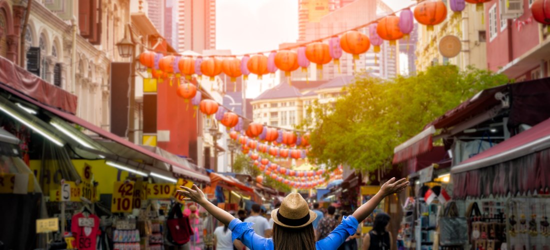 Blick auf eine belebte Marktstraße in Singapur bei Sonnenuntergang. Eine Frau mit Strohhut und Rucksack streckt freudig die Arme in die Höhe. Die Straße ist gesäumt von bunten Läden mit Verkaufsständen und über ihr hängen viele rote Lampions. Im Hintergru