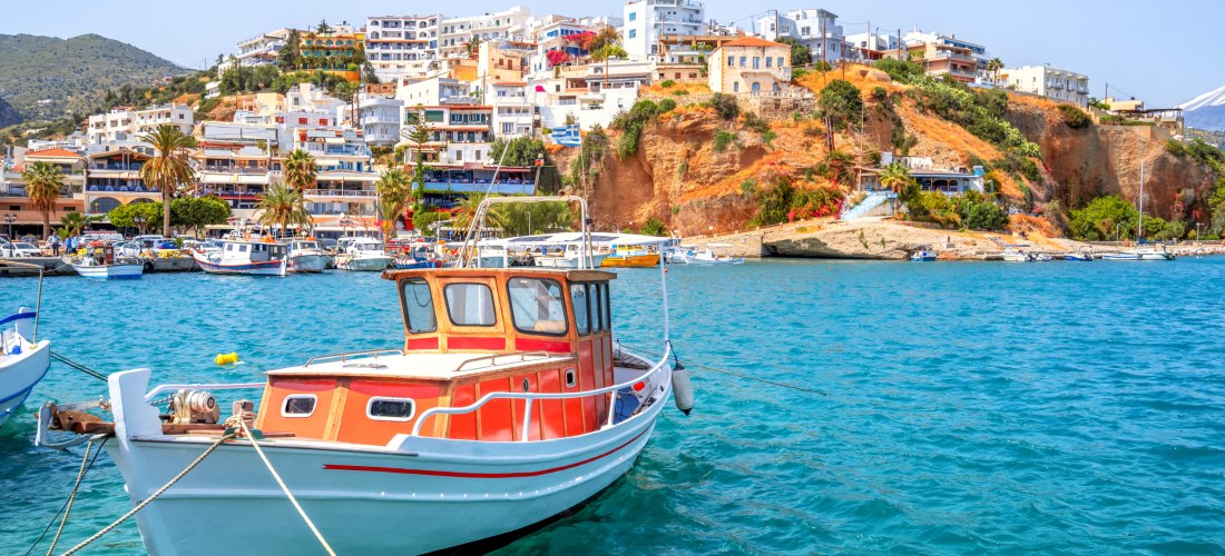 Ein traditionelles Holzboot liegt im türkisfarbenen Wasser eines kleinen Hafens auf Kreta. Im Hintergrund erstreckt sich eine bunte Küstenstadt mit weiß getünchten Häusern, Hotels und Palmen, die sich an einen Hügel schmiegen. Die Sonne scheint hell, der 