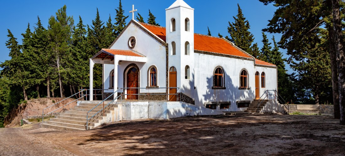 Kleine weiß getünchte Kapelle mit rotem Ziegeldach und Glockenturm, gelegen an einem Waldweg. Die Kirche ist von Bäumen umgeben und strahlt in der Nachmittagssonne