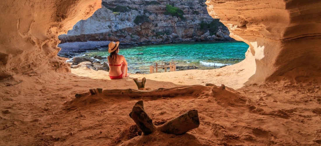 Eine Frau in einem roten Bikini und Strohhut sitzt in einer natürlichen Sandsteinhöhle und blickt aufs türkisfarbene Meer hinaus. Die Höhle öffnet sich zu einer kleinen, geschützten Bucht mit Felsen und einem Holzsteg. Die Szene vermittelt Ruhe und Naturv