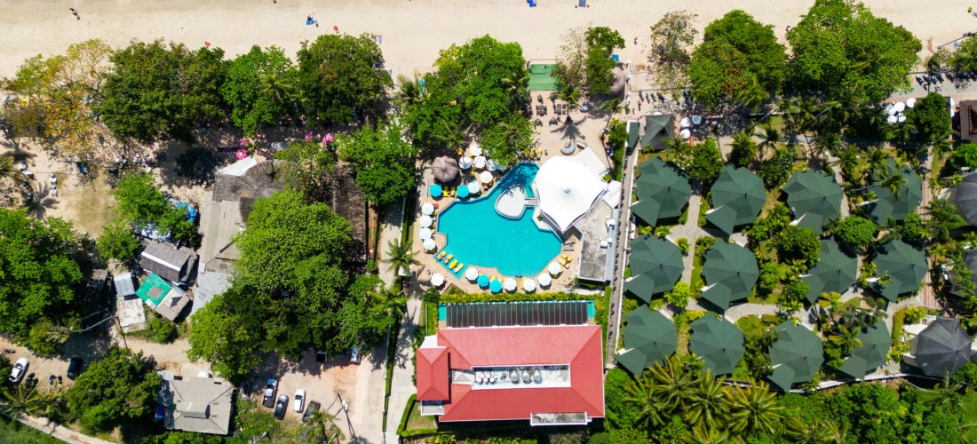 Das Bild zeigt eine senkrechte Luftaufnahme eines tropischen Strandresorts in Thailand. Im Zentrum befindet sich ein großer, organisch geformter Swimmingpool mit hellblauen Liegen und weißen Sonnenschirmen ringsherum. Direkt dahinter erstreckt sich ein he