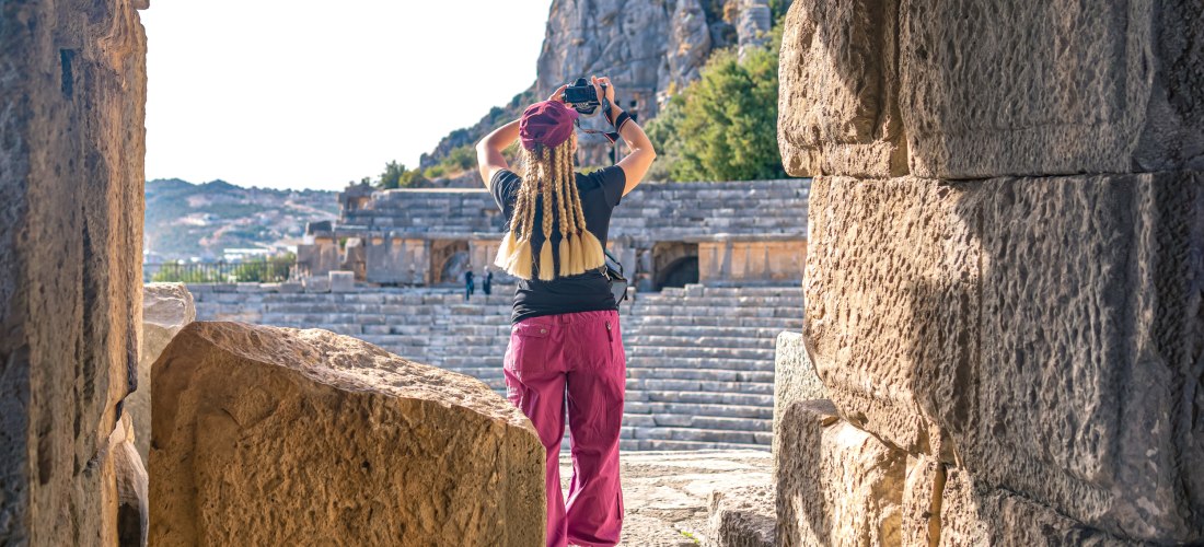 Eine Person mit langen, geflochtenen Haaren fotografiert ein antikes Amphitheater. Sie steht in einem steinernen Torbogen, im Hintergrund sind Stufen und Felsen zu sehen