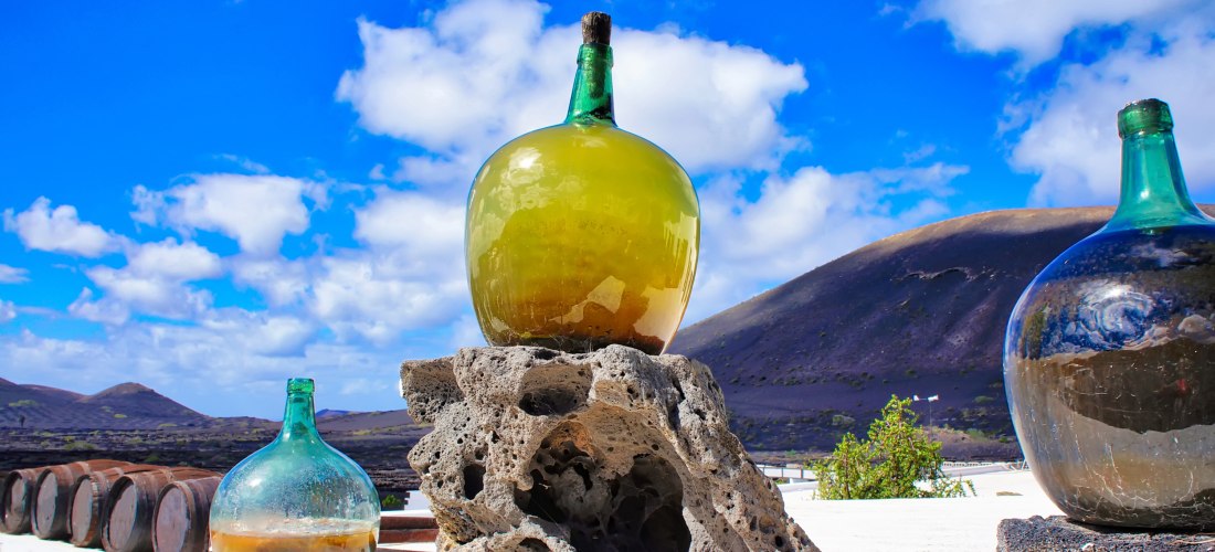 Große grüne Glasflasche, die auf einem Lava-Steinblock steht, vor einem blauen Himmel und vulkanischen Hügeln im Hintergrund. Weitere Flaschen und Holzfässer sind sichtbar.