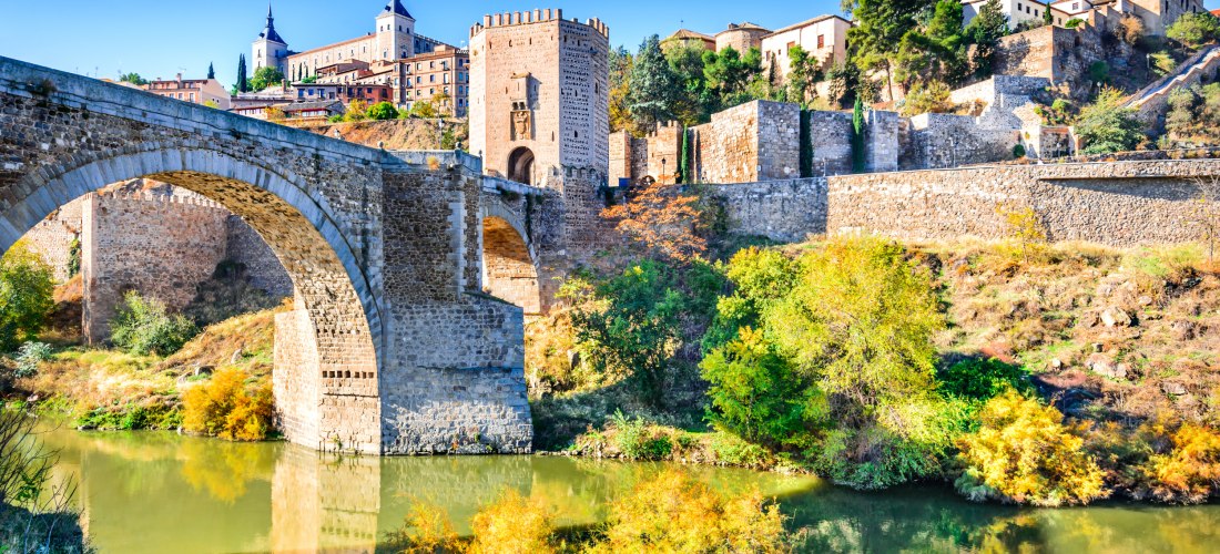 Steinerne Brücke Puente de San Martín über den Fluss Tajo in Toledo, Spanien. Im Hintergrund ist die mittelalterliche Altstadt mit dem Alcázar von Toledo zu sehen. Herbstliche Bäume spiegeln sich im ruhigen Wasser.