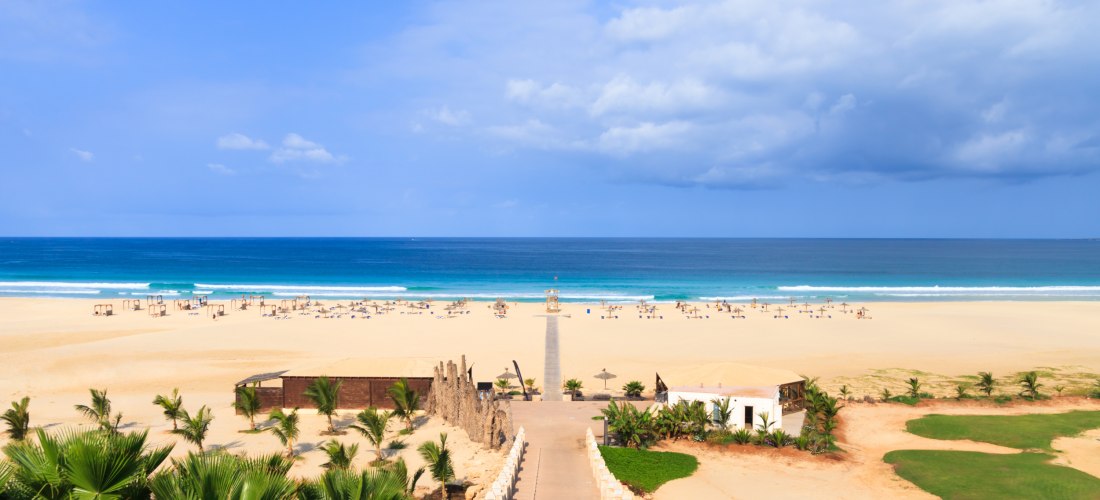 Breiter, heller Sandstrand auf der kapverdischen Insel Boa Vista mit Blick auf das tiefblaue Meer. Sonnenliegen und Schirme sind über den Strand verteilt, ein gerader Weg führt von einem Hotelgelände mit Palmen durch den Sand bis ans Wasser. Der Himmel is