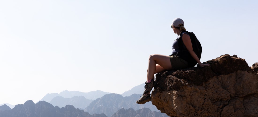 Wanderin mit Rucksack sitzt auf einem Felsvorsprung und blickt auf eine Berglandschaft