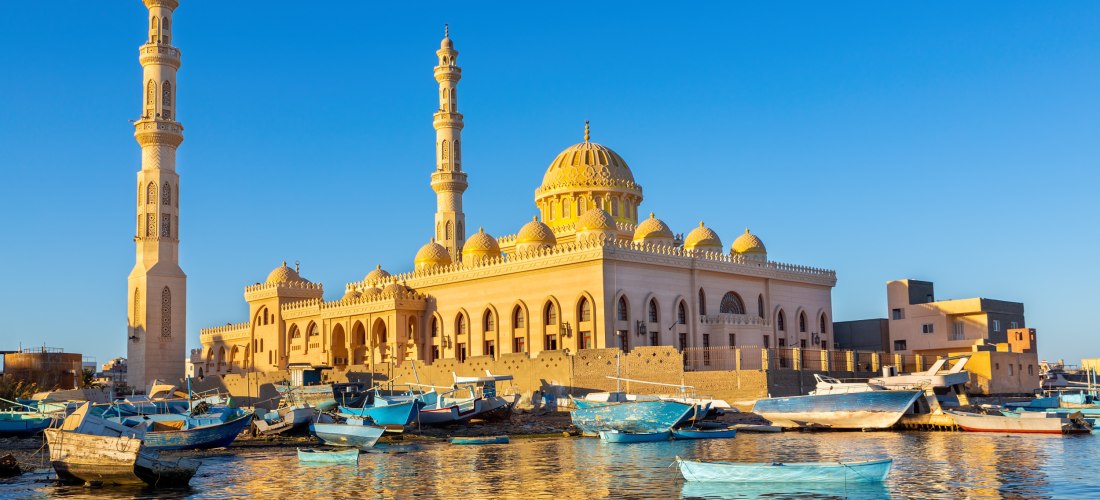 Im warmen Licht der untergehenden Sonne erstrahlt eine eindrucksvolle Moschee mit filigraner Architektur, goldener Kuppel und zwei reich verzierten Minaretten. Sie liegt direkt am Wasser, wo viele bunte Holzboote vor Anker liegen und sich sanft im ruhigen
