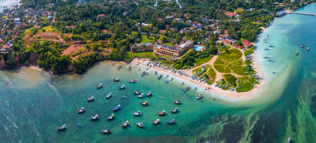 Luftaufnahme einer tropischen Küstenstadt mit roten Dächern, Fischerbooten im türkisfarbenen Wasser und kleinen Buchten. Der Ort liegt auf einer Halbinsel, umgeben von grünem Land, bunten Häusern und klarem Meer. Zahlreiche Boote ankern in der Bucht. Die 