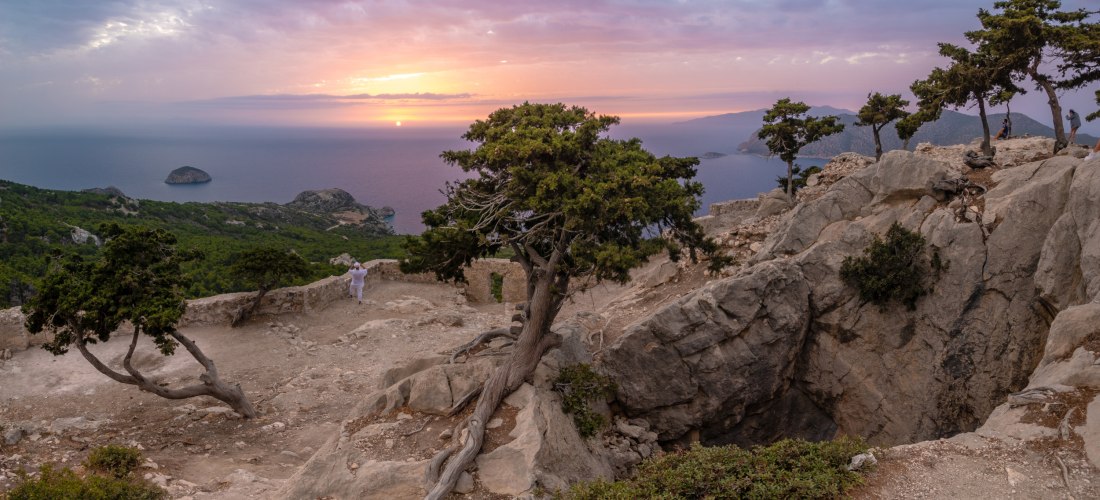 Panoramablick von einem felsigen Bergplateau auf das Meer bei Sonnenuntergang. Im Vordergrund wachsen knorrige Bäume zwischen großen Felsblöcken. Eine Person in Weiß fotografiert die Szene. Die Sonne geht am Horizont unter, der Himmel ist rosa-violett gef