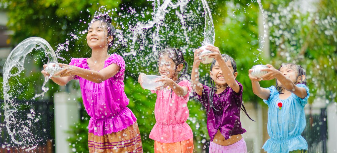 Vier lachende Kinder in bunten traditionellen Kleidern werfen Wasser mit kleinen Schalen in die Luft. Sie feiern ausgelassen im Freien, umgeben von grünen Bäumen