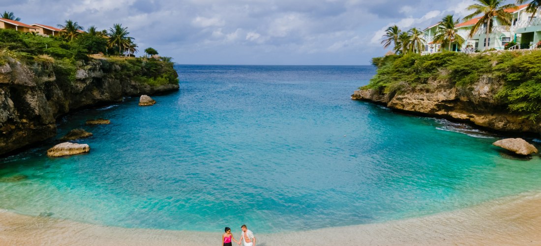 Kleine, geschützte Bucht auf Curaçao mit klarem türkisblauem Wasser. Zwei Personen gehen am hellen Sandstrand entlang. Links und rechts säumen grüne Klippen mit Palmen und Häusern den Eingang zur Bucht, darüber ein teils bewölkter Himmel.