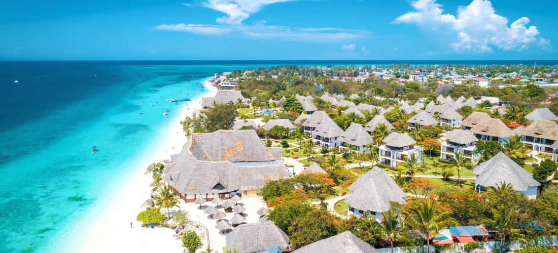 Luftaufnahme eines tropischen Strandresorts mit strohgedeckten Bungalows, Palmen, weißem Sandstrand und langem türkisblauem Küstenabschnitt.