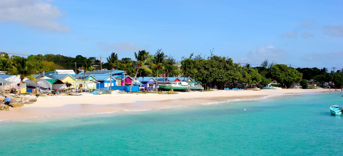 Fischerdorf Oistins: Küstenansicht auf Barbados mit türkisfarbenem Wasser, weißem Sandstrand und einer Reihe bunter, kleiner Häuser und Boote am Ufer. Palmen und grüne Bäume im Hintergrund.