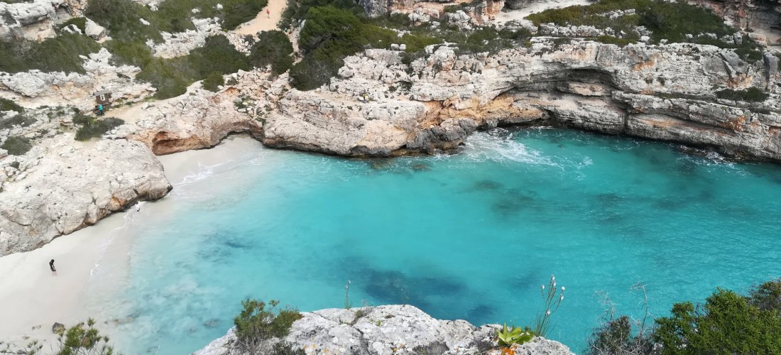 Eine versteckte Bucht mit türkisfarbenem Wasser, umgeben von hohen, zerklüfteten Felswänden auf Mallorca. Eine kleine sandige Bucht liegt eingebettet zwischen den Felsen, der Strand ist fast menschenleer. Ein schmaler Pfad führt durch die grüne Vegetation