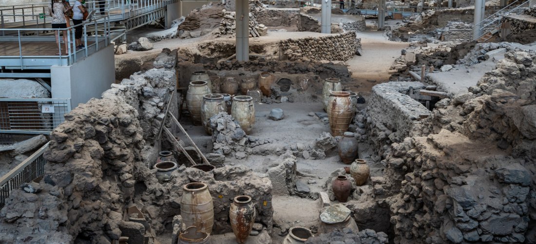 Archäologische Ausgrabungsstätte von Akrotiri auf Santorini. Im Bild sind antike, aus Ton gefertigte Vorratsgefässe zwischen den freigelegten Ruinen von Steingebäuden zu sehen. Die Szenerie ist überdacht und mit Stegen und Plattformen ausgestattet, auf de