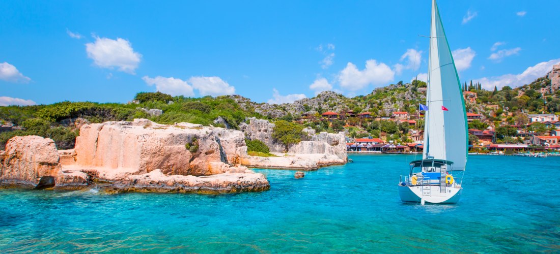 Ein weißes Segelboot fährt über türkisblaues Wasser entlang einer felsigen Küste mit grüner Vegetation. Im Hintergrund liegt ein malerisches Dorf mit traditionellen Häusern am Hang, unter strahlend blauem Himmel.