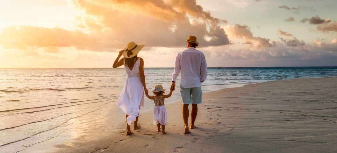 Familie spaziert bei Sonnenuntergang barfuß am Strand entlang, mit Blick aufs Meer.