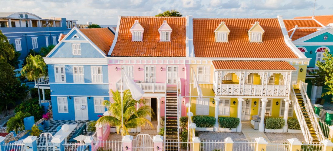 Reihe farbenfroher Kolonialhäuser in Willemstad auf Curaçao. Die Gebäude sind in leuchtendem Blau, Rosa und Gelb gestrichen und haben rote Ziegeldächer. Die Architektur ist typisch niederländisch-karibisch, mit Balkonen und weißen Fensterläden
