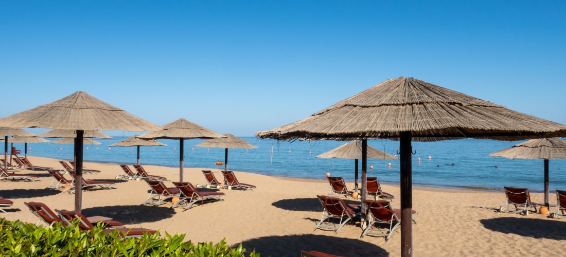 Sonnenliegen und Strohdach-Sonnenschirme an einem Sandstrand in Fujairah, Vereinigte Arabische Emirate. Perfekter Ort zum Entspannen und Sonnenbaden am Meer.