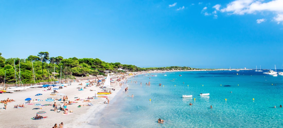 Breiter, feinsandiger Strand auf Formentera mit zahlreichen Menschen, die am Strand liegen oder im klaren, türkisblauen Wasser baden. Links im Bild stehen viele bunte Sonnenschirme und Boote liegen am Rand des Strandes. Im Hintergrund erstreckt sich ein d