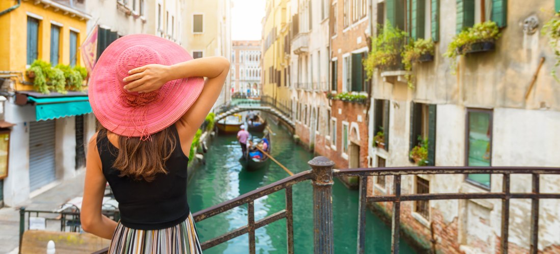 Eine Frau mit einem pinkfarbenen Sonnenhut steht auf einer kleinen Brücke in Venedig und blickt auf einen Kanal mit Gondeln. Die umliegenden Häuser sind alt, bunt und mit Fensterläden und Blumenkästen geschmückt. Die Szene wirkt sommerlich und romantisch.