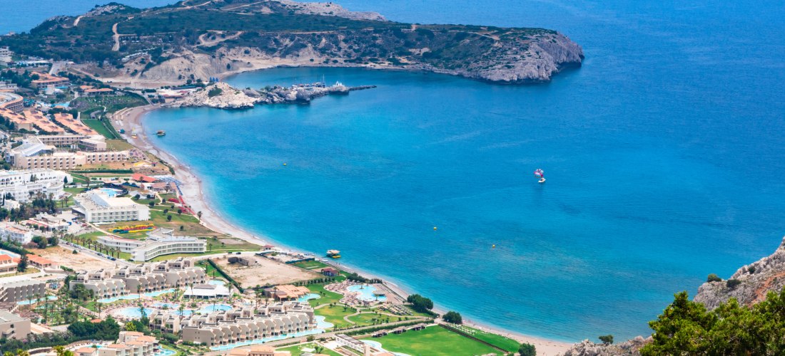 Das Bild zeigt eine weite Bucht mit türkisblauem Meer auf einer griechischen Insel – wahrscheinlich Rhodos.    Am langen, flach abfallenden Sandstrand reihen sich große Hotelanlagen mit Pools, Gärten und Sportflächen.   Links im Bild erkennt man eine klei