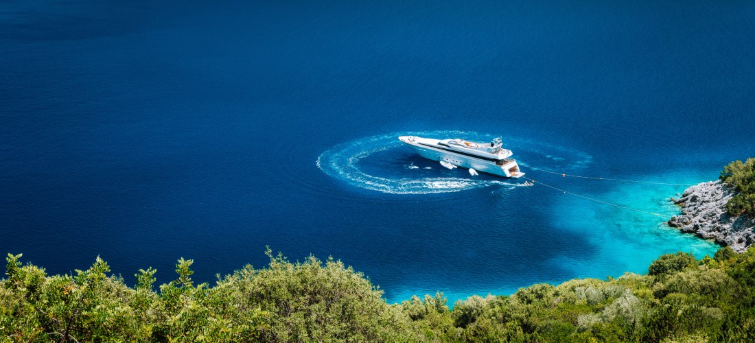 Exklusiver Moment auf dem Wasser: Eine elegante Luxusyacht fährt durch eine smaragdgrüne Bucht, das türkisfarbene Wasser wirbelt in sanften Kreisen um den Rumpf. Die Szene wird eingerahmt von grüner Vegetation und felsiger Küste – mediterraner Luxus pur.