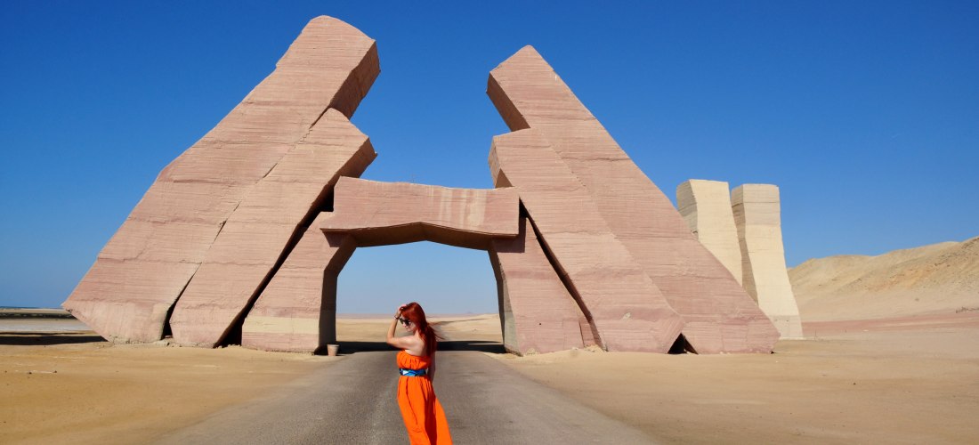 Eine Frau mit langen roten Haaren und einem leuchtend orangefarbenen Kleid steht barfuß auf einer Straße mitten in der Wüste. Im Hintergrund erhebt sich ein monumentales, modernes Steintor aus mehreren schräg stehenden, kantigen Blöcken in rötlichen Farbt