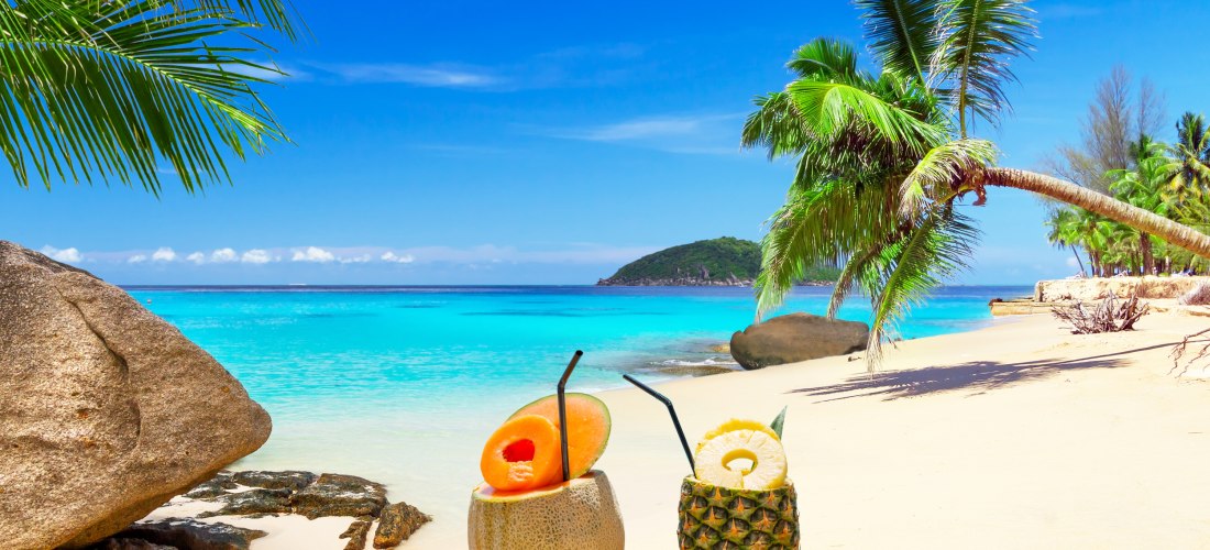 Zwei tropische Cocktails in einer Melone und einer Ananas am weißen Sandstrand mit Palmen und türkisblauem Meer
