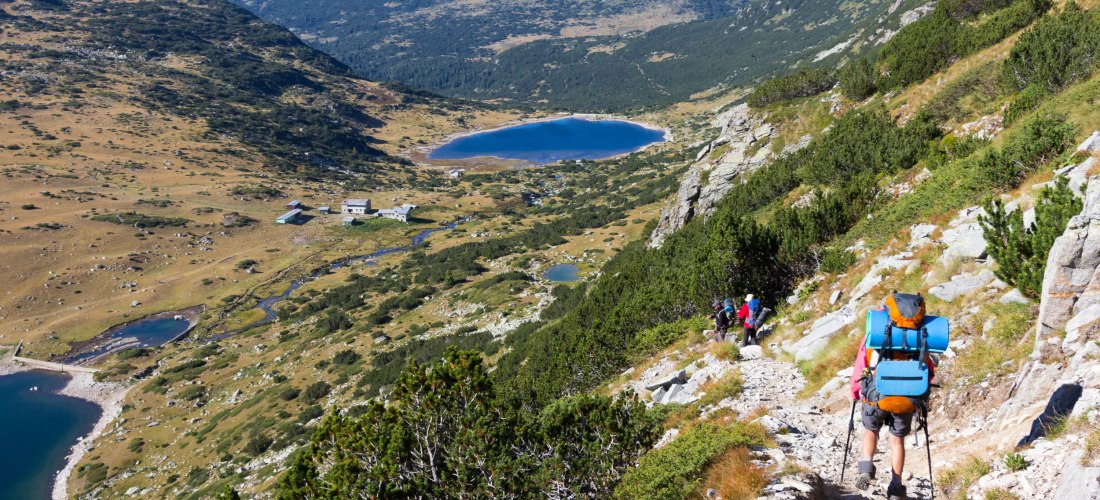 Wanderer mit Rucksack und Trekkingstöcken auf einem steinigen Bergpfad. Der Weg führt durch das grüne Rila-Gebirge in Bulgarien. Im Hintergrund liegen mehrere klare, blaue Bergseen eingebettet in eine weite, offene Landschaft mit sanften Hügeln und verstr