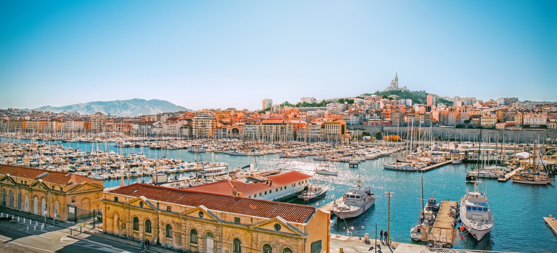 Vieux-Port von Marseille mit vielen Segelbooten, terrakottafarbenen Häusern und der Basilika Notre-Dame de la Garde auf dem Hügel unter klarem Himmel.